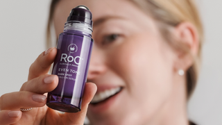 A close-up lifestyle shot of a woman holding a purple rollerball bottle of ROC Even Tone Dark Spot Cell Turnover Serum, with a soft-focus background of her face.