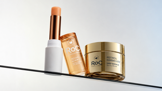 Three OTC skincare products from ROC Paris arranged on a black shelf against a light gray background: a white stick with a peach-tan cap, an amber/orange bottle, and a gold jar labeled Retinol Correction Deep Wrinkle Eye Cream.