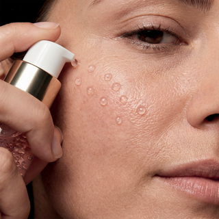 Close-up of a person applying a clear gel or serum from a pump bottle onto their cheek, with visible droplets on the skin.