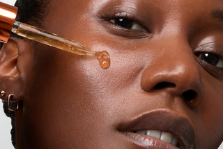 Close-up of a person's face with a glass dropper applying amber-colored serum onto the cheek, showing a glossy drop of the product.