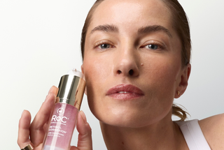 A close-up lifestyle shot of a woman holding a bottle of R.O.C. Natural Correction serum against a clean white background.