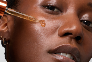 Close-up of a person's face with a gold-toned dropper applying a golden serum onto the cheek, showing a glossy finish.