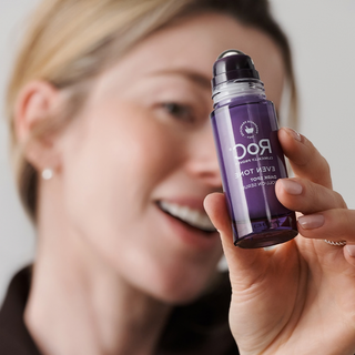 A woman holds a purple rollerball bottle of Rod's Even Toner against a soft, blurred background.