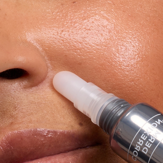 Close-up of a skincare rollerball applicator being pressed onto a person's cheek, showing a clear silicone roller tip and part of a dark metallic tube with white text.