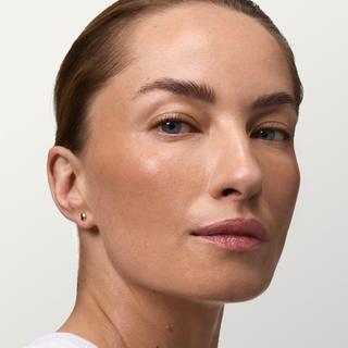 Close-up portrait of a woman wearing a small gold stud earring against a light gray background.