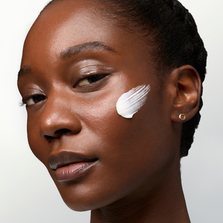 Close-up of a woman's face with a thick swipe of white cream or balm applied to her cheek against a plain light background.