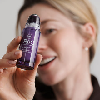 A woman holds up a purple roll-on bottle of Roa Laundry Mending Even Toner, with a soft-focus background of her face.