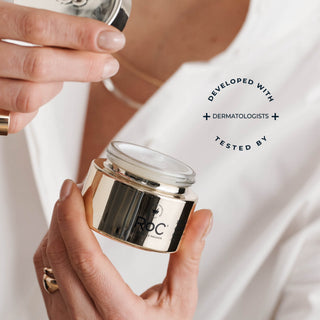 A person in a white top holds a small cosmetic jar with a reflective gold-toned label and clear lid. Another hand holds a matching container above.